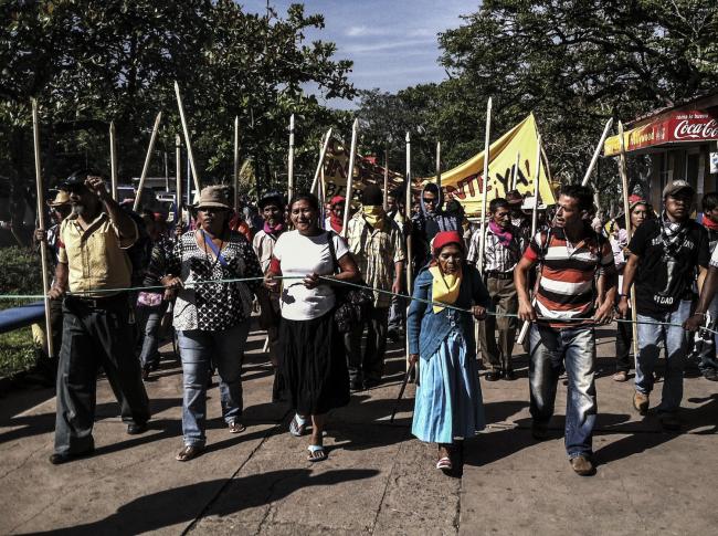 Vásquez, center, leads march to Presidential Palace to demand a just trial for Berta Cáceres' assassination, Tegucigalpa. The poles, representing authority, are used by Lenca elders. (Photo by Beverly Bell) 