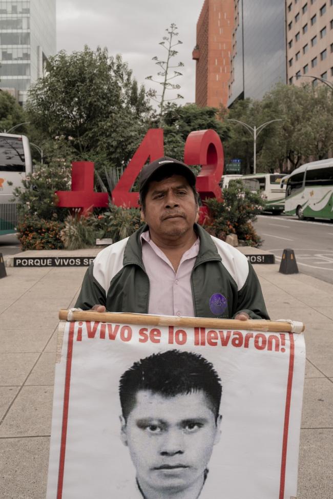 Francisco Lauro Villegas, a father of one of the missing Ayotzinapa students, attends the march holding a poster of his missing child. The poster features the phrase 